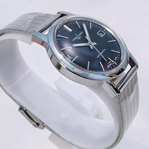 May include: A silver-toned Ulysse Nardin automatic watch with a dark blue face and a date window at the 3 o'clock position. The watch has a mesh band and is displayed on a white stand. The watch face has the text "AUTOMATIC" and "SWISS MADE".