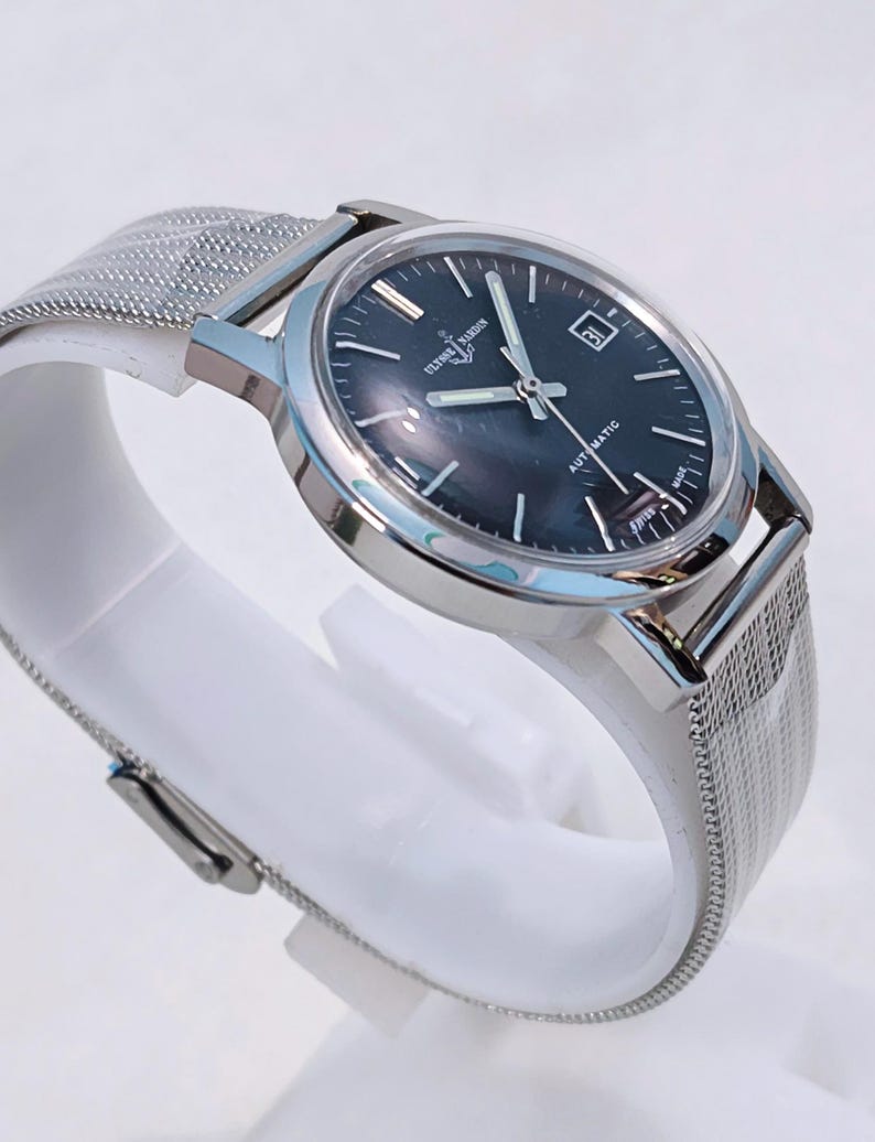 May include: A silver-toned Ulysse Nardin automatic watch with a dark blue face and a date window at the 3 o'clock position. The watch has a mesh band and is displayed on a white stand. The watch face has the text "AUTOMATIC" and "SWISS MADE".