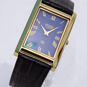 May include: A rectangular gold-toned Seiko Quartz watch with a blue face and gold Roman numerals. The watch has a black leather strap. The watch face displays the Seiko Quartz logo. A classic watch design.