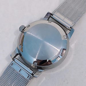 May include: Close-up of a silver wristwatch with a mesh band. The watch case is round and polished, with a crown on the side. The band is made of a fine, woven metal. The watch is shown against a light background.