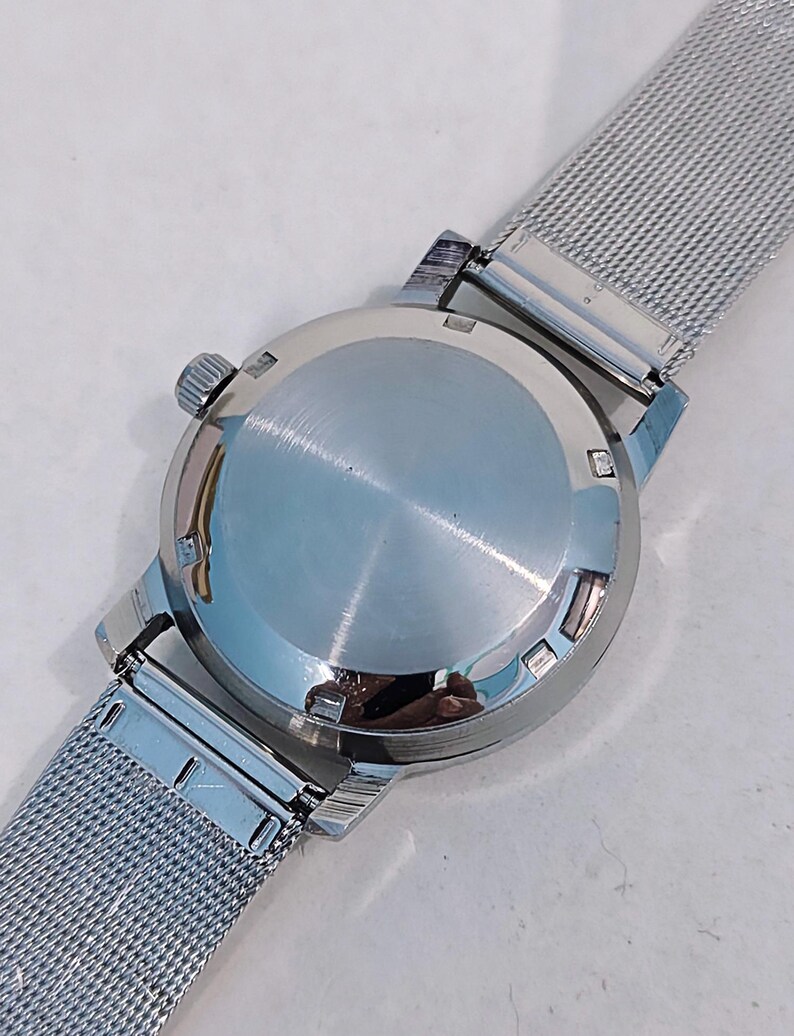 May include: Close-up of a silver wristwatch with a mesh band. The watch case is round and polished, with a crown on the side. The band is made of a fine, woven metal. The watch is shown against a light background.