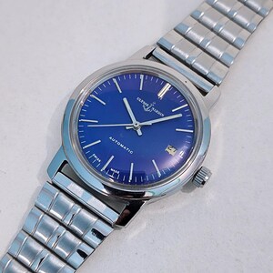 May include: A silver-toned Ulysse Nardin automatic wristwatch with a vibrant blue dial. The watch features silver hands and markers, a date window, and a stainless steel band. The watch face has the text "Ulysse Nardin" and "Automatic".