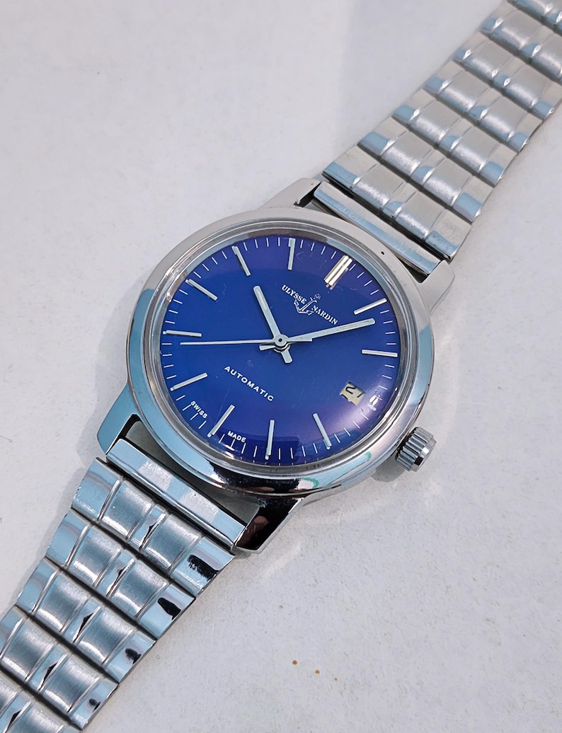 May include: A silver-toned Ulysse Nardin automatic wristwatch with a vibrant blue dial. The watch features silver hands and markers, a date window, and a stainless steel band. The watch face has the text "Ulysse Nardin" and "Automatic".