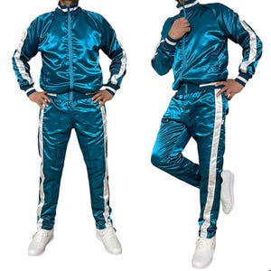Könnte beinhalten: Türkisfarbener Satin-Trainingsanzug mit weißen Streifen. Die Jacke hat einen Reißverschluss und einen Stehkragen. Die Hose hat einen Kordelzugbund und ein gerades Bein. Getragen mit weißen Turnschuhen.