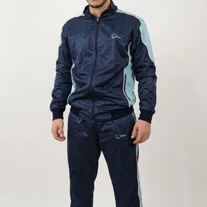 Könnte beinhalten: Marineblauer Trainingsanzug mit hellblauen Akzenten. Die Jacke hat einen durchgehenden Reißverschluss und das Wort "Dmw" ist auf der Brust geschrieben. Die Hose hat einen passenden hellblauen Streifen an der Seite. Der Trainingsanzug ist aus glänzendem Material.