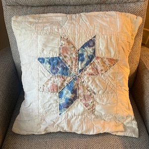 May include: A square, cream-coloured cushion featuring a quilted star pattern. The star's points are crafted from floral fabric in shades of pink, blue, and purple. The cushion rests on a grey upholstered chair.