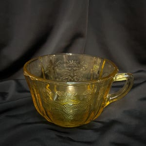 May include: A yellow glass teacup with a floral design. The cup has a rounded shape and a handle.