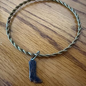 May include: A bronze-colored twisted wire bracelet with a small, dark bronze cowboy boot charm. The bracelet is circular and rests on a wooden surface, showcasing a rustic, country-western theme.
