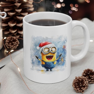 Op de afbeelding: Witte keramische mok met een aquarel-illustratie van een minion die een kerstmanmuts draagt en zijn armen in een vrolijke pose uitstrekt. De minion staat in een besneeuwde scène met een blauwe achtergrond.