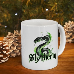 May include: White ceramic mug with a green snake and the words "Slytherin" in black lettering. The snake is surrounded by green watercolor-like splatters.