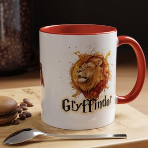 Op de afbeelding: Witte keramische mok met een rode rand en handvat. De mok heeft een aquarel-illustratie van een leeuw met een Gryffindor-wapen en de tekst "Gryffindor".
