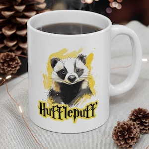 Op de afbeelding: Witte keramische mok met een gele en zwarte dassenillustratie en de tekst "Hufflepuff" in zwarte letters.