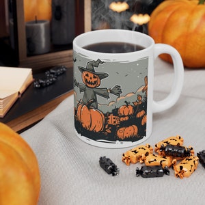 Op de afbeelding: Een witte keramische mok met een Halloween-thema ontwerp met een pompoenkop-vogelverschrikker die in een veld met pompoenen staat. De mok heeft een zwart handvat en is gevuld met koffie.
