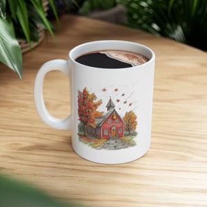 May include: White ceramic mug featuring a red and yellow illustration of a small red building with a bell tower, surrounded by trees with orange and yellow leaves. The building is set against a background of a light grey sky with rain and falling leaves.
