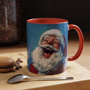 Op de afbeelding: Een rode en blauwe keramische koffiemok met een feestelijk beeld van een lachende kerstman. De mok heeft een rode rand en een rode handgreep.