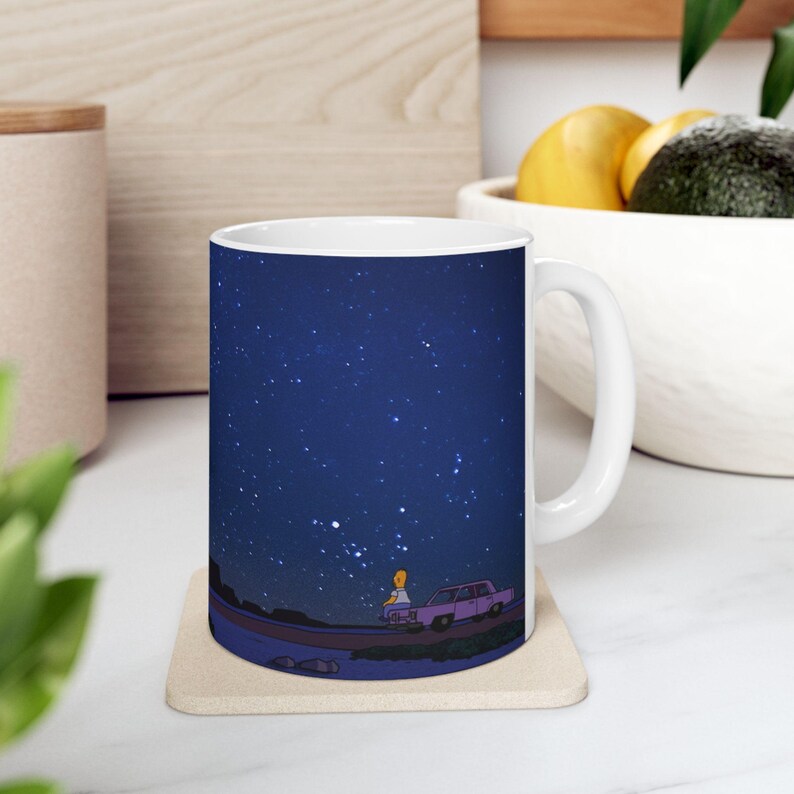 K&ouml;nnte beinhalten: Wei&szlig;e Keramiktasse mit einem bedruckten Design eines blauen Nachthimmels mit Sternen. Eine Comicfigur steht im Vordergrund neben einem Auto.