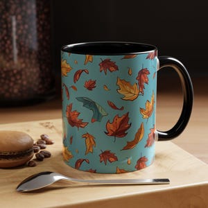 Könnte beinhalten: Eine blaue Keramiktasse mit schwarzem Rand und Henkel. Die Tasse zeigt ein Muster aus bunten Herbstblättern auf hellblauem Hintergrund.