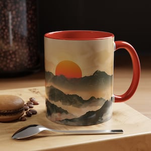 May include: A red and white ceramic mug with a sunset over mountains design. The mug has a red rim and handle.
