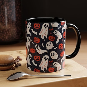 Op de afbeelding: Zwarte keramische mok met een Halloween-thema. De mok heeft een patroon van witte geesten en oranje pompoenen op een donkerblauwe achtergrond.