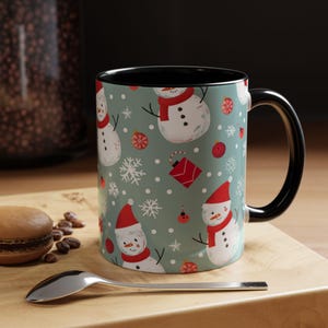 Peut inclure: Une tasse en céramique noire et blanche avec un motif de bonhomme de neige de Noël. La tasse a une anse noire et un fond bleu clair avec des bonhommes de neige rouges, blancs et noirs.