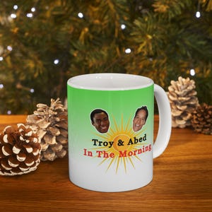 Puede incluir: Taza de cerámica blanca con un fondo degradado de verde a blanco. La taza presenta un sol de dibujos animados con las caras de Troy y Abed de la serie de televisión Community. El texto "Troy & Abed In The Morning" está impreso debajo del sol.