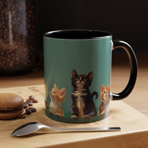 Op de afbeelding: Een teal keramische mok met een zwarte rand en handvat heeft een afbeelding van vier kittens die omhoog kijken. De kittens zijn een zwart-witte tabby, een ginger tabby en twee oranje tabbies.