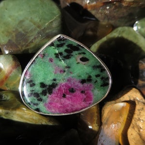 May include: A green and pink gemstone ring with black flecks set in a silver band. The ring is photographed on a bed of smooth, colorful stones.