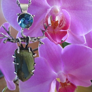 May include: A silver pendant with a green and blue gemstone, a silver tree branch design, and a blue topaz stone. The pendant is displayed on a pink orchid.