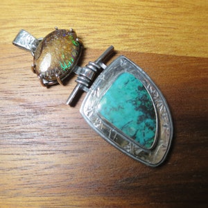 May include: Two silver pendants on a wooden surface. One pendant features a brown and iridescent stone, while the other has a turquoise stone set in a decorative silver frame. The pendants are connected by a silver bar.