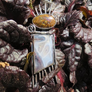 May include: A silver pendant necklace with a brown and iridescent opal stone set in a silver frame. The pendant is surrounded by silver spikes and hangs from a silver chain. The pendant is resting on a bed of dark purple leaves.