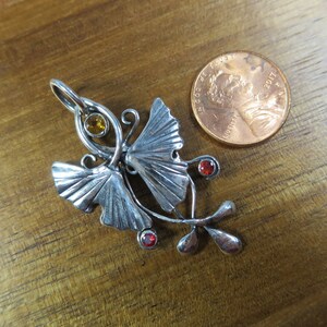 Puede incluir: Pendiente de plata con dos hojas de ginkgo estilizadas con una piedra amarilla y dos piedras rojas. El pendiente es aproximadamente del tama&ntilde;o de una moneda de un centavo de euro.