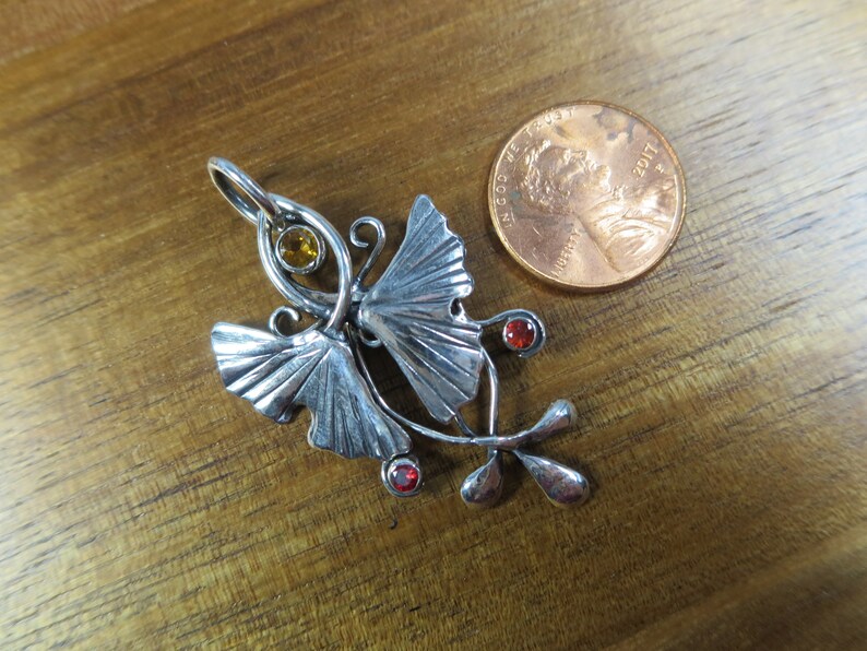 Puede incluir: Pendiente de plata con dos hojas de ginkgo estilizadas con una piedra amarilla y dos piedras rojas. El pendiente es aproximadamente del tama&ntilde;o de una moneda de un centavo de euro.