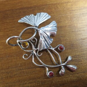 Puede incluir: Un colgante de plata con dos hojas de ginkgo estilizadas y un dise&ntilde;o delicado y en espiral. El colgante est&aacute; adornado con tres piedras preciosas rojas y una piedra preciosa amarilla.