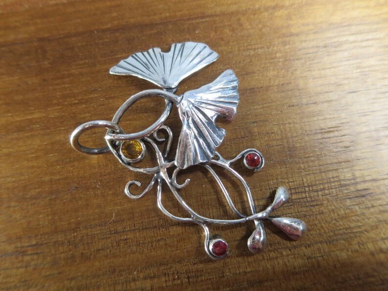 Puede incluir: Un colgante de plata con dos hojas de ginkgo estilizadas y un dise&ntilde;o delicado y en espiral. El colgante est&aacute; adornado con tres piedras preciosas rojas y una piedra preciosa amarilla.