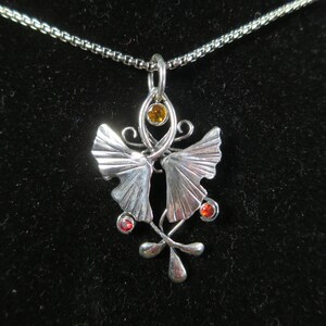 Puede incluir: Un collar de plata con un colgante que presenta dos hojas de ginkgo estilizadas con una piedra amarilla y dos piedras rojas.