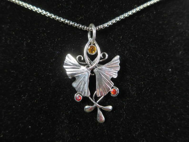 Puede incluir: Un collar de plata con un colgante que presenta dos hojas de ginkgo estilizadas con una piedra amarilla y dos piedras rojas.