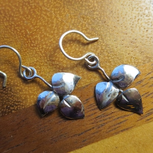 May include: Silver-toned leaf earrings. Each earring features three stylized leaves with a textured surface, connected to a simple hook. The earrings are displayed against a warm, wooden background.