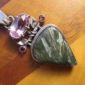 May include: A silver pendant with a green and white stone, a large pink oval stone, and two smaller pink stones. The pendant is on a wooden surface.