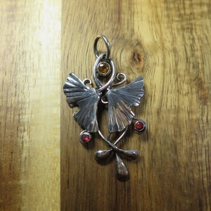 Puede incluir: Pendiente de plata con dos hojas de ginkgo estilizadas con piedras preciosas rojas y naranjas.