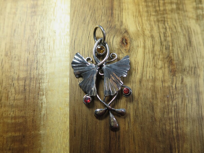Puede incluir: Pendiente de plata con dos hojas de ginkgo estilizadas con piedras preciosas rojas y naranjas.