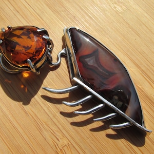 May include: A silver pendant with a brown agate stone and a teardrop-shaped amber stone. The pendant has a geometric design with a series of prongs extending from the agate stone.