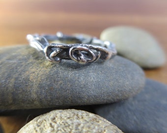 Könnte beinhalten: Ein silberner Ring mit einem einzigartigen, organischen Design, das an einen verdrehten Ast oder eine Rebe erinnert. Der Ring liegt auf einem Hintergrund aus grauen Steinen.