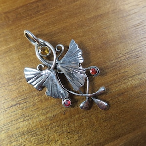 Puede incluir: Un colgante de plata con dos hojas de ginkgo estilizadas con piedras preciosas rojas y amarillas. El colgante tiene un dise&ntilde;o delicado e intrincado, perfecto para agregar un toque de naturaleza a cualquier atuendo.