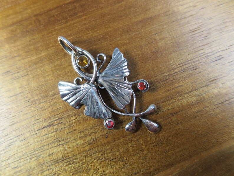 Puede incluir: Un colgante de plata con dos hojas de ginkgo estilizadas con piedras preciosas rojas y amarillas. El colgante tiene un dise&ntilde;o delicado e intrincado, perfecto para agregar un toque de naturaleza a cualquier atuendo.