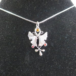 Puede incluir: Un collar de plata con un colgante de mariposa con piedras preciosas rojas y naranjas. El colgante est&aacute; decorado con dise&ntilde;os intrincados y una cadena delicada.