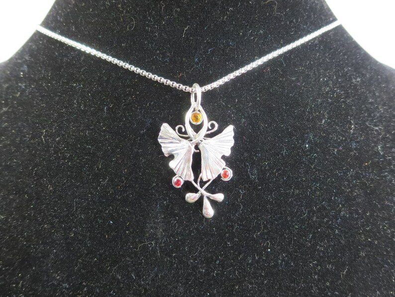 Puede incluir: Un collar de plata con un colgante de mariposa con piedras preciosas rojas y naranjas. El colgante est&aacute; decorado con dise&ntilde;os intrincados y una cadena delicada.
