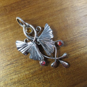 Puede incluir: Un colgante de plata con dos hojas de ginkgo estilizadas, una peque&ntilde;a piedra preciosa &aacute;mbar en el centro y dos piedras preciosas rojas en el tallo.