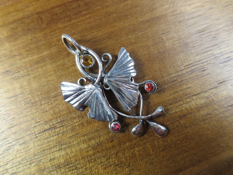Puede incluir: Un colgante de plata con dos hojas de ginkgo estilizadas, una peque&ntilde;a piedra preciosa &aacute;mbar en el centro y dos piedras preciosas rojas en el tallo.