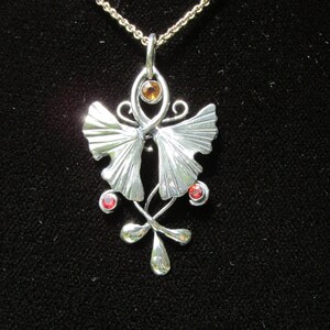 Puede incluir: Un collar con un colgante de plata con un dise&ntilde;o estilizado de hoja de ginkgo. El colgante presenta dos hojas con una peque&ntilde;a piedra preciosa naranja en el centro y dos piedras preciosas rojas en la parte inferior.