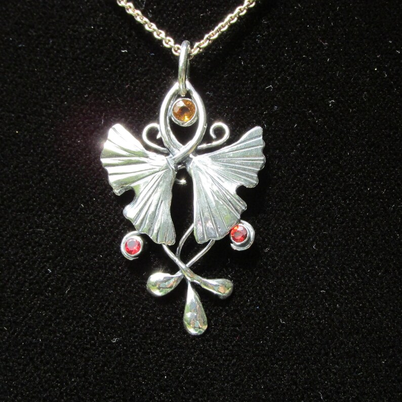 Puede incluir: Un collar con un colgante de plata con un dise&ntilde;o estilizado de hoja de ginkgo. El colgante presenta dos hojas con una peque&ntilde;a piedra preciosa naranja en el centro y dos piedras preciosas rojas en la parte inferior.